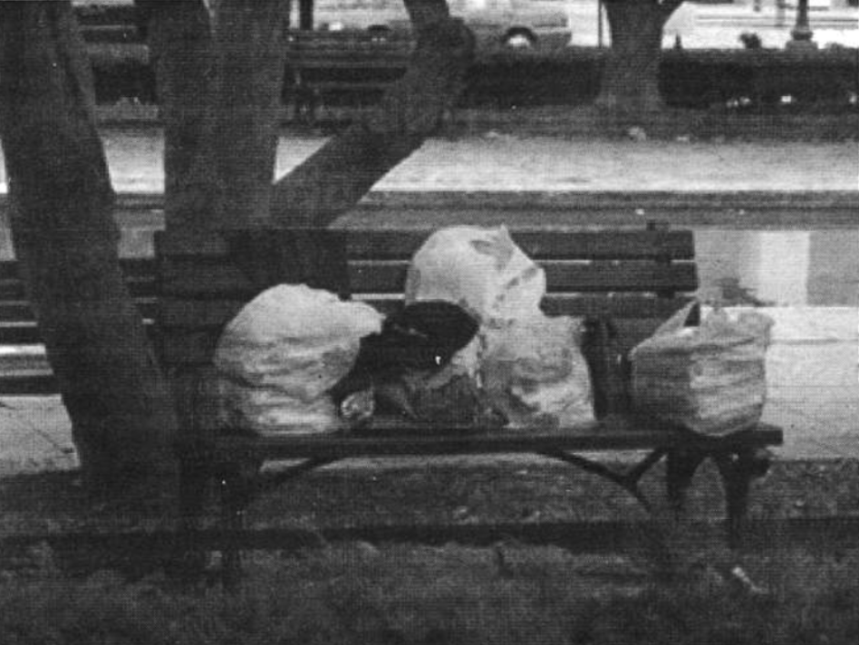 A black and white photo shows multiple trash bags that contain someone's belongings sitting on a park bench.