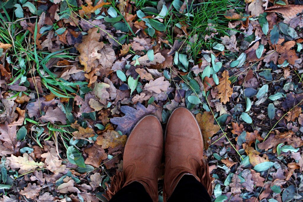 Photo of someone's shoes as they are standing outside. There is green grass and old fallen tree leaves.