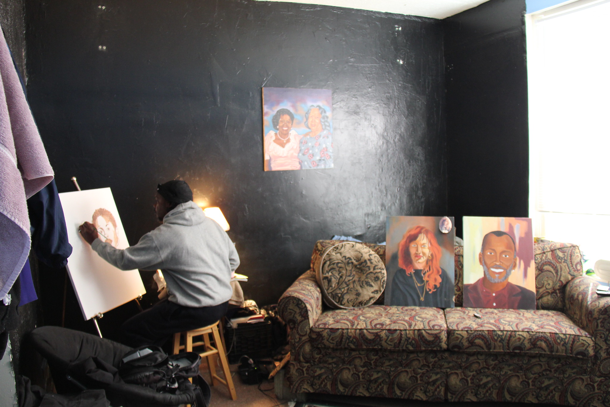 Photo showing Reggie Dakari painting with his back to the camera. Two other paintings sit on a nearby couch and a third hangs on the wall.