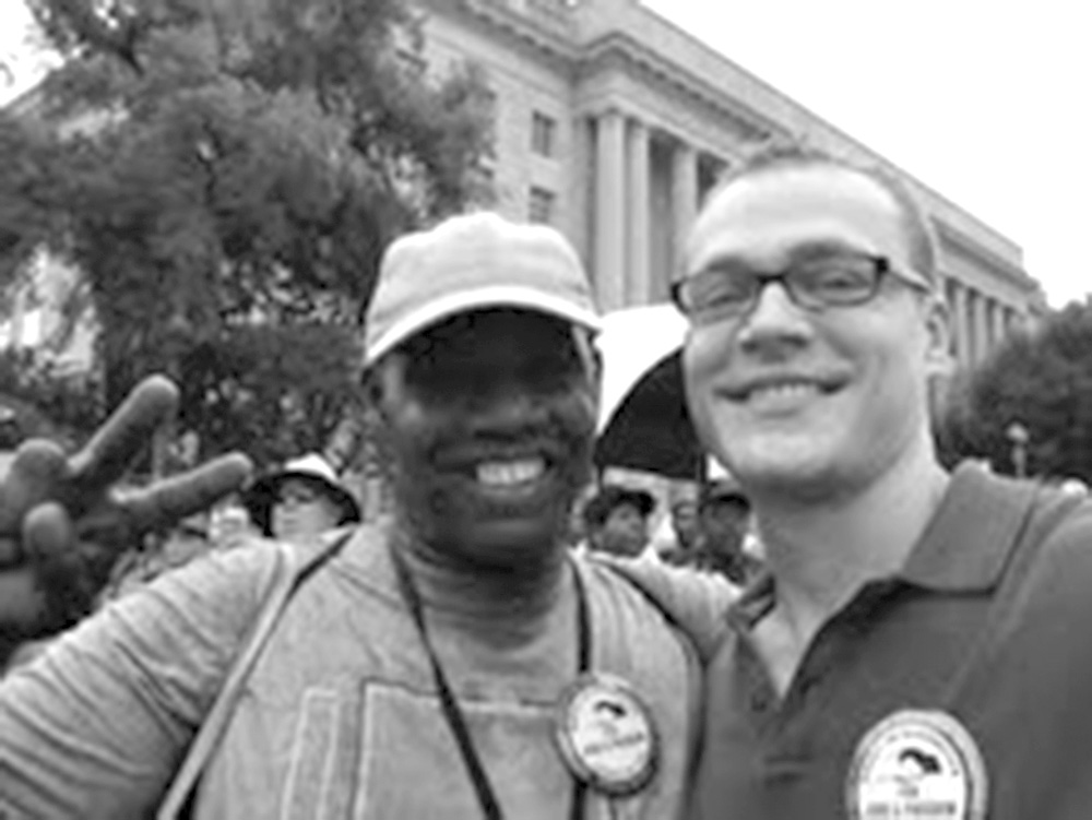 Black and white of two men standing together and smiling. They are both wearing a matching button from the anniversary event.