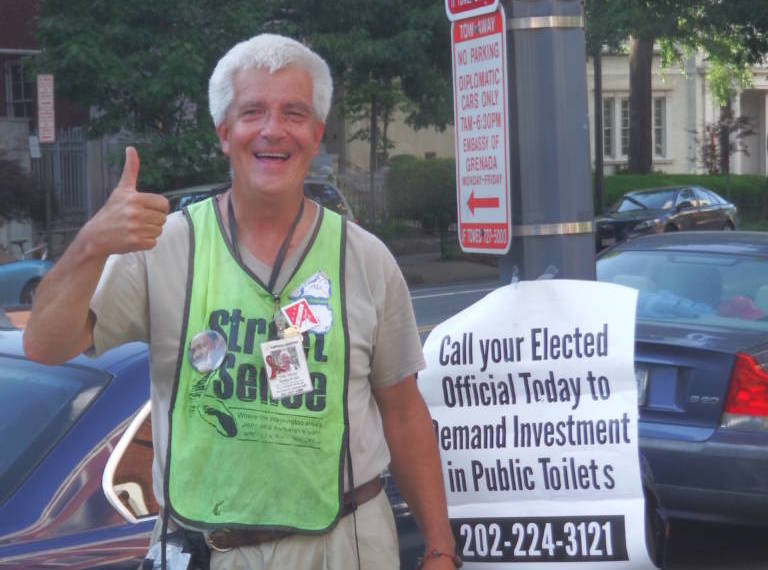 Photo of Gary Minter in downtown D.C., wearing a "Street Sense" badge and vest, standing next to a sign that says "Call your Elected Official Today to Demand Investment in Public Toilets: 202-224-3121" and giving a thumbs up.