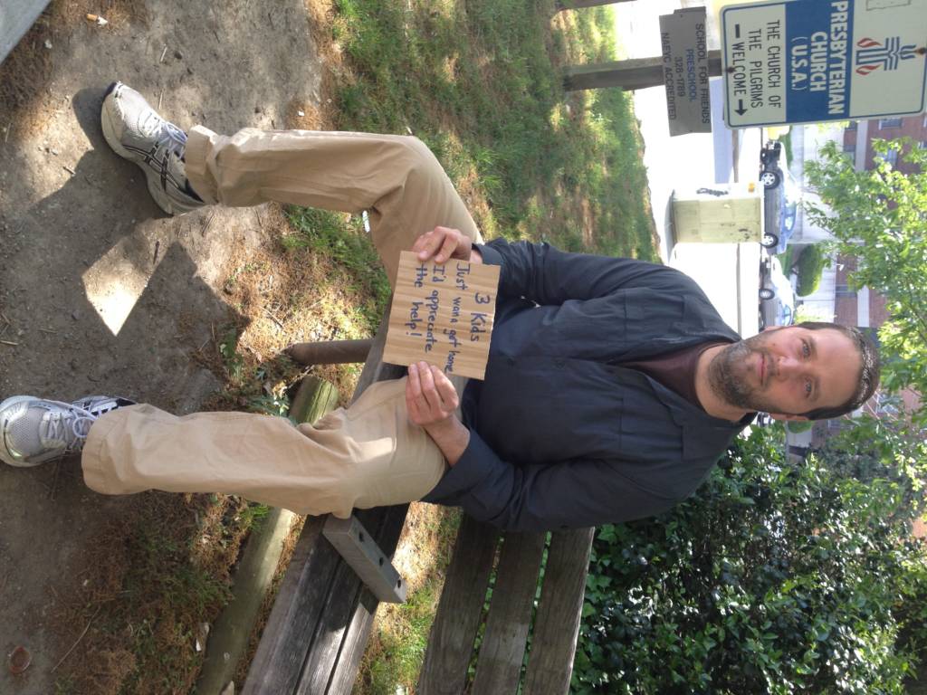 CEO Eric Weires sits on a bench and displays his sign. Sign reads: “3 Kids. Just wana get home. I’d Appreciate the help!”