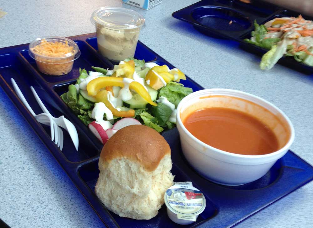 A blue hard plastic lunch tray holding a roll, soup and a salad, sitting on top of a cafeteria table.