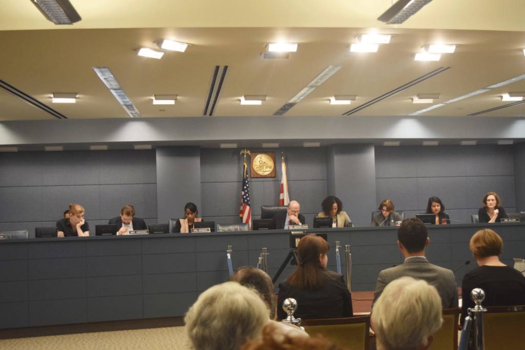 Photo of several people sitting at a table to testify in front of the councilmembers seated at the dais facing them. The heads of a few people seated to observe the hearing are also visible.