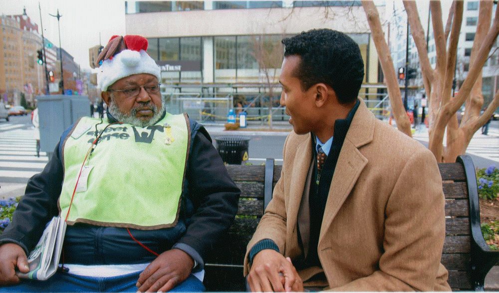 Photo of Anthony wearing a Street Sense vest and Santa hat, sitting next to a man in a nice tan coat.