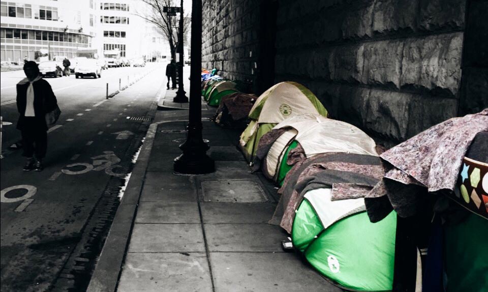 Photo showing a line of tents down the sidewalk under an underpass near Union Station. The photo is black and white, showing only the tents in color. A pedestrian walks in the street instead of near the tents.