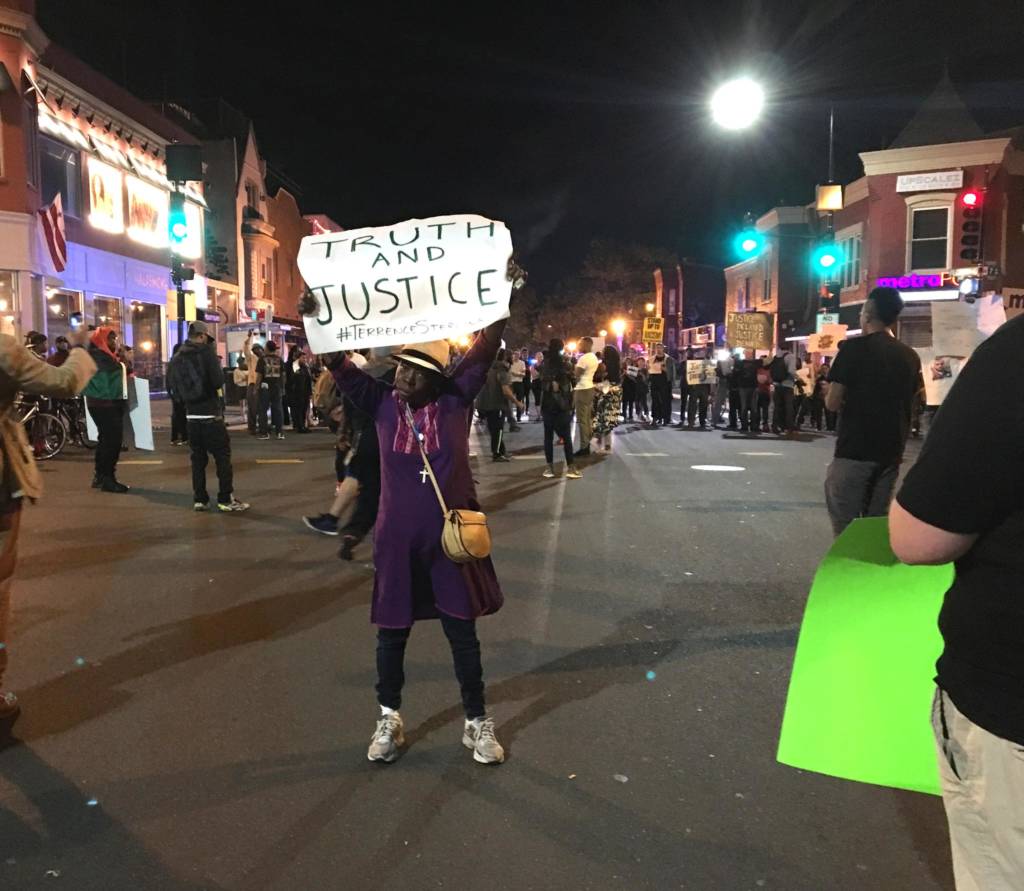 An older Black woman stand in the middle of the street holding a sign that reads "Justice" above her head