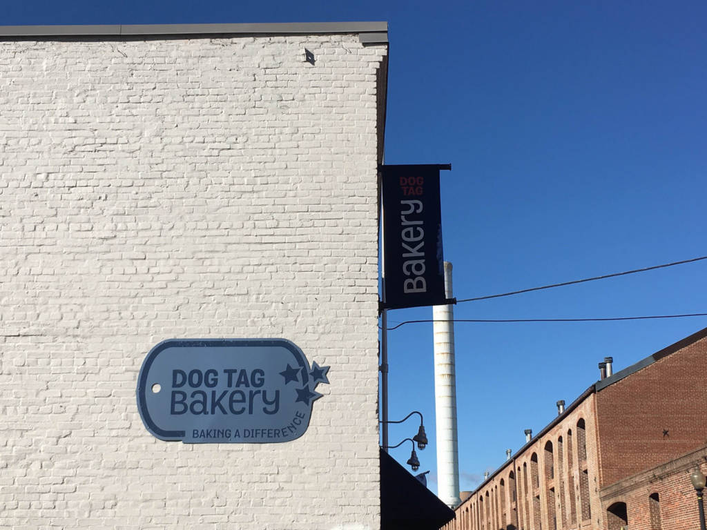 A grey brick building with a sign in the shape of a dog tag reading: "Dog Tag Bakery."
