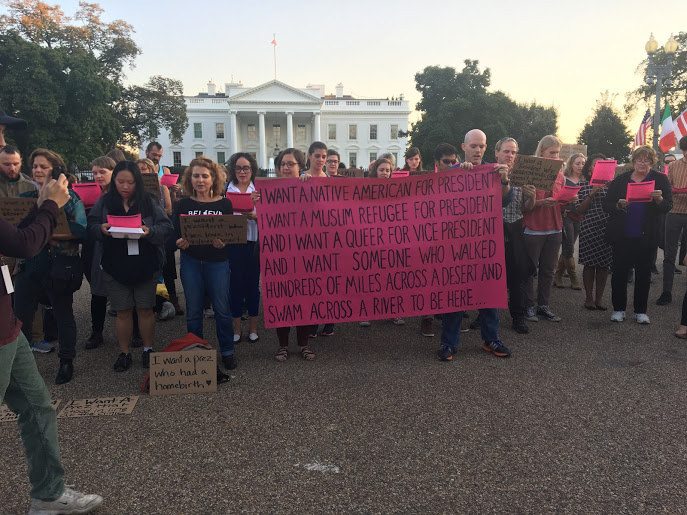 A large group of people hold signs and read from papers in front of the White House. In front, two people hold a large pink banner reading: "I WANT A NATIVE AMERICAN FOR PRESIDENT. I WANT A MUSLIM FOR PRESIDENT. AND I WANT A QUEER FOR VICE PRESIDENT. AND I WANT SOMEONE WHO WALKED HUNDREDS OF MILES ACROSS A DESERT AND SWAM ACROSS A RIVER TO BE HERE..."