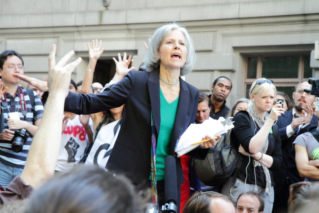 Jill Stein, a woman with grey hair, a green shirt and black blazer, gives a speech to a large crowd of people around her. She is holding a clipboard and pencil in one hand and is gesturing outwards with the other.