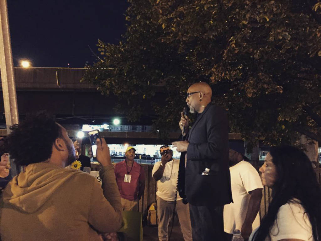 Ajamu Baraka stands speaking into a microphone in front of a crowd at night.
