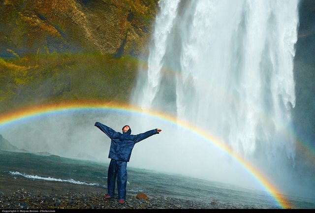 Man standing under rainbow in front of waterfall