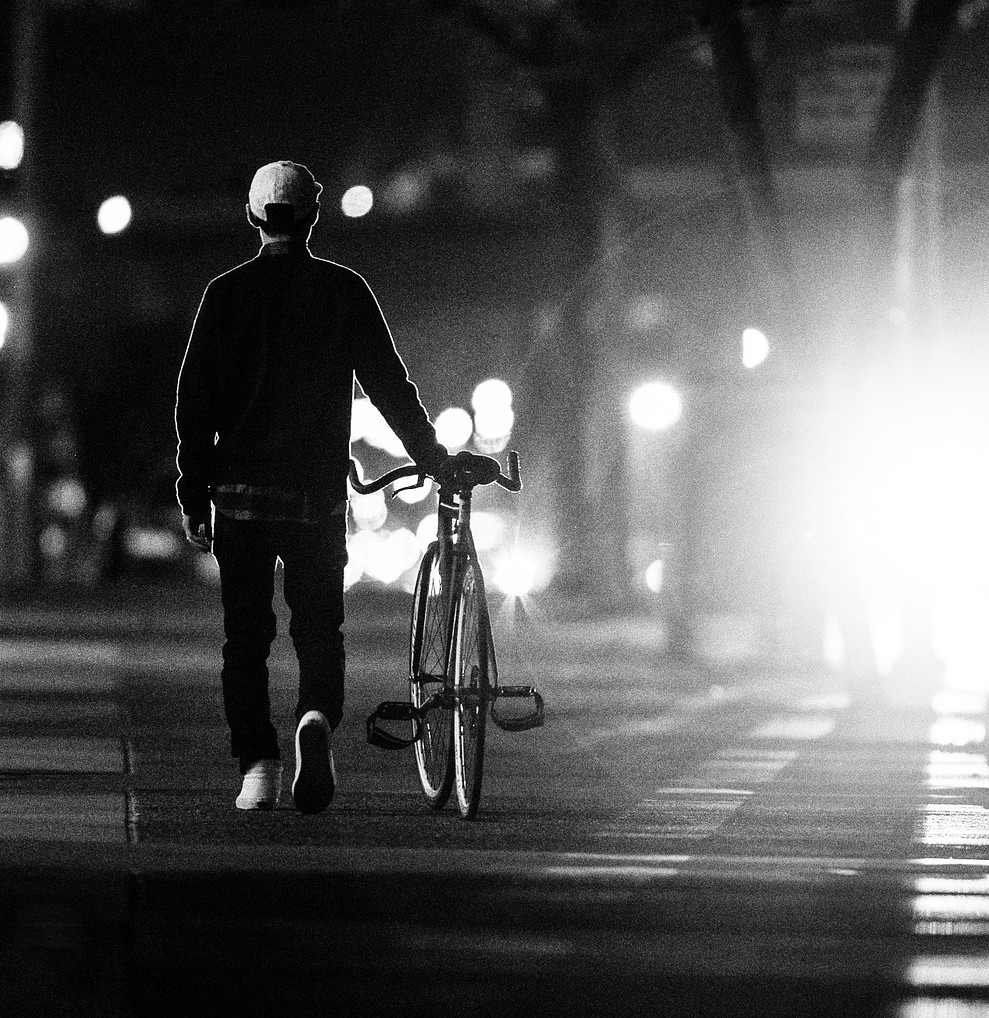 Man walking his bike on a city street with lights in the background.