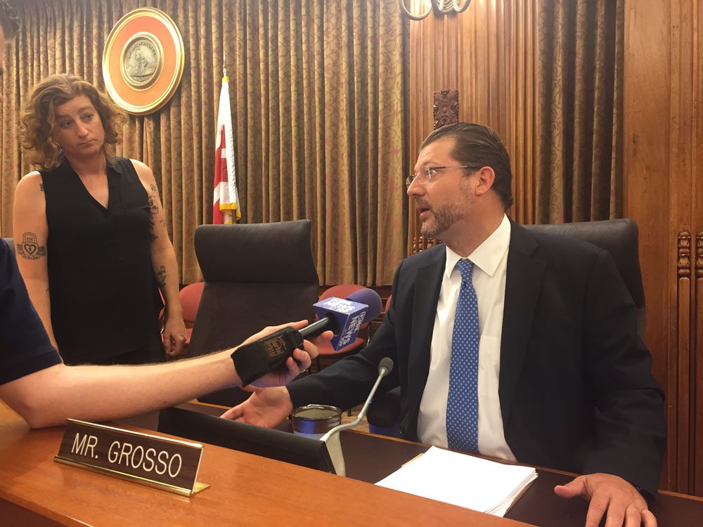 A photo of Councilmember Grosso answering media questions from the D.C. Council Dais, flanked by staffer Darby Hickey.