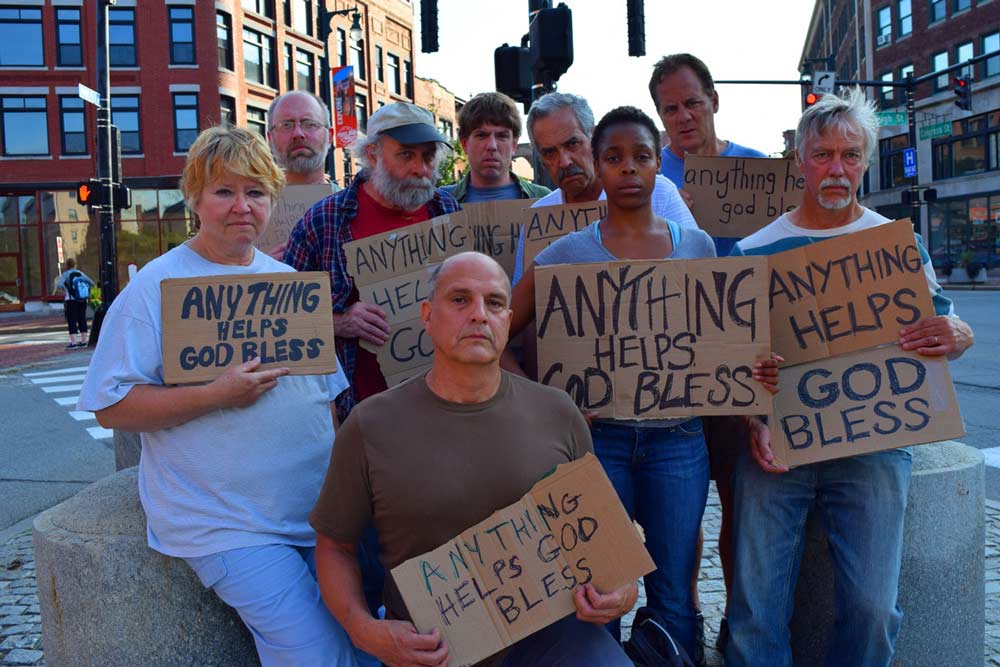 A group of people on a city street, all facing the camera, each holding cardboard signs that state "Every Thing Helps God Bless"