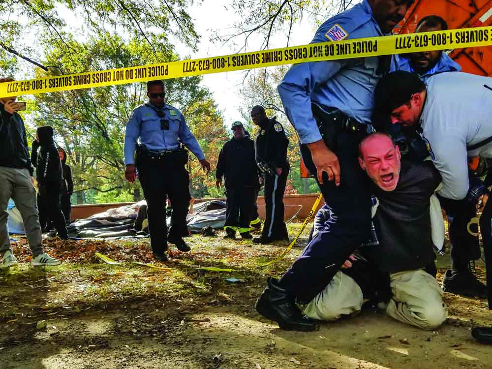 A photo showing 3 officers restraining a man, who is on his knees screaming, as others look on. A police tape line runs in front of them and the orange of a city dump truck is visible behind them.