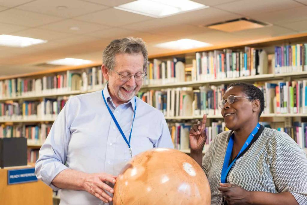 Dr. Jim Green, standing, holds a globe of Mars and points out some of the geography to Cynthia Mewborn. A row of books in the library at the NASA headquarters is behind them.