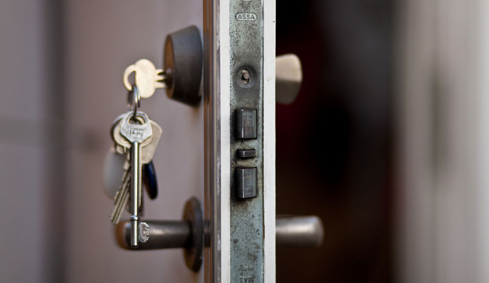 Photo of an open door with keys hanging out of the lock. The background inside the doorway is blurred.