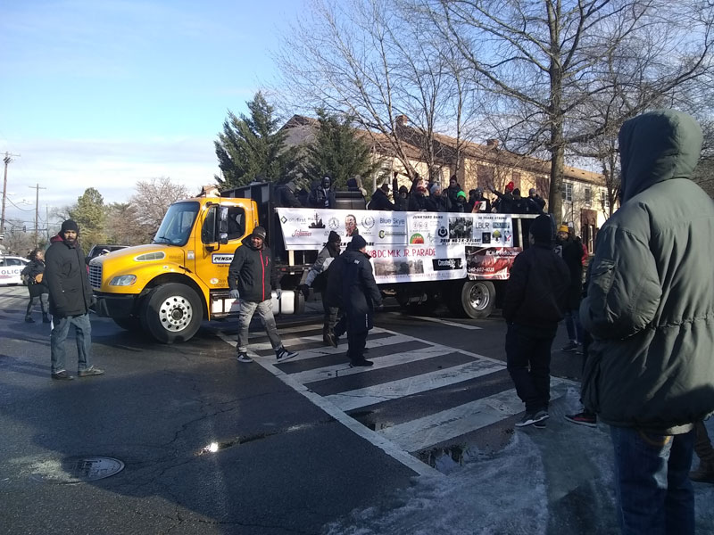 Photo of a truck holding councilmembers, other speakers and supports as it drives through a street with a few men standing around it.