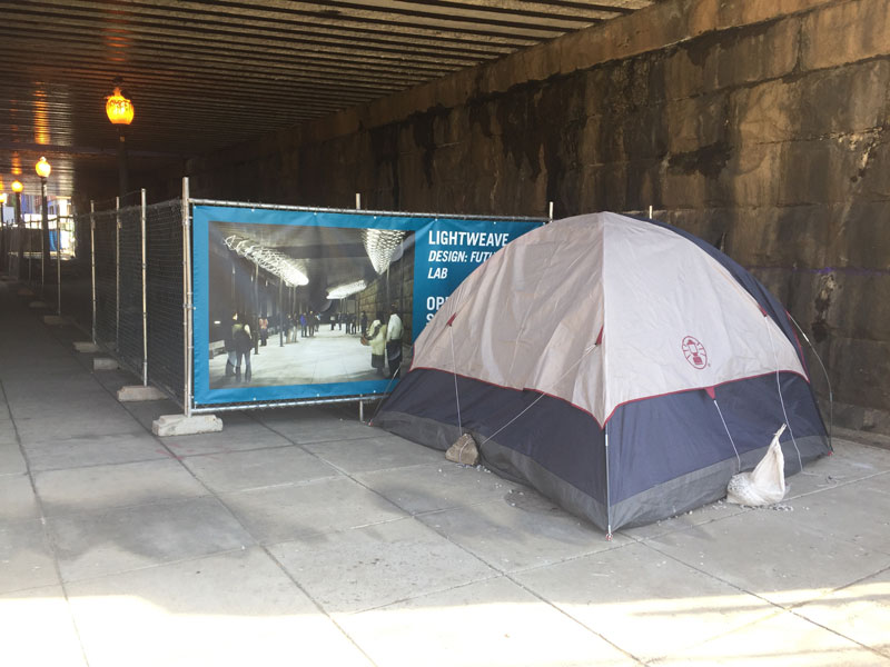 Photo of a tent in an underpass, right next to a fence with a poster advertising the new light installations.