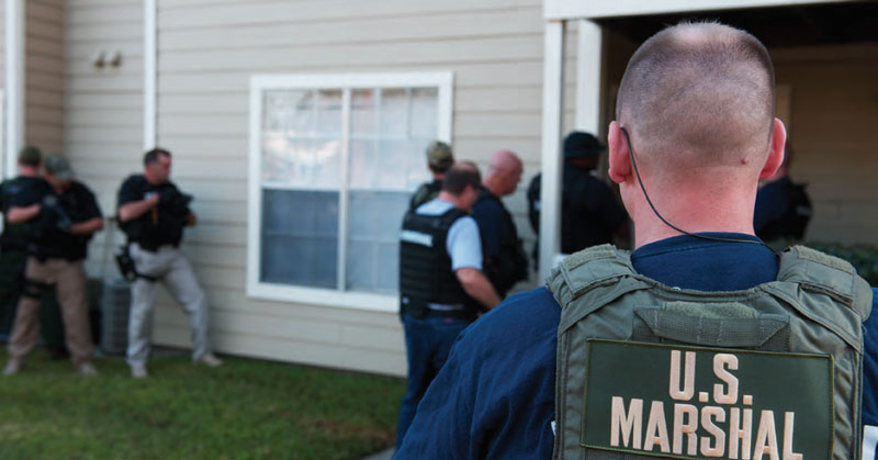 Photo of the back of a U.S. Marshal facing a home while other officers stand around the house in the background.