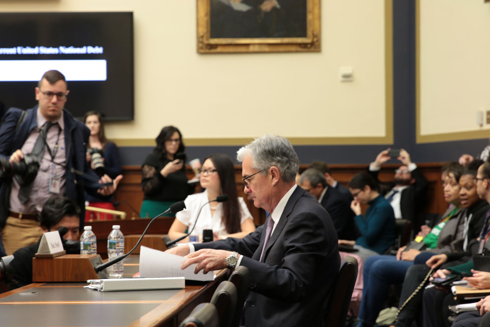 Profile view of Jerome Powell at a desk, flipping through a report as he answers questions from Congress. He is surrounded by photographers and observers of the hearing.
