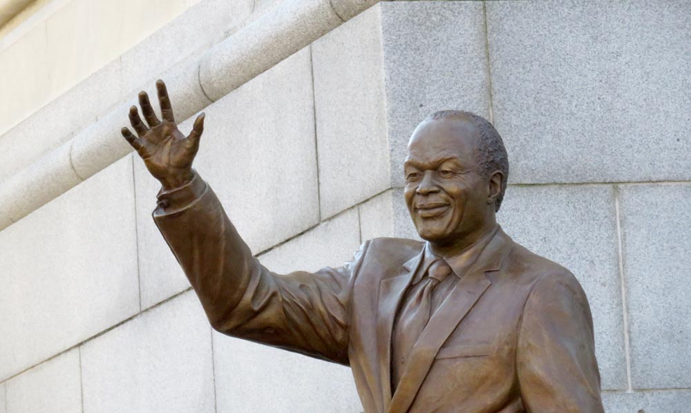 Photo of the upper half of a bronze statue of Marion Barry wearing a suit, smiling and raising his right arm to wave.