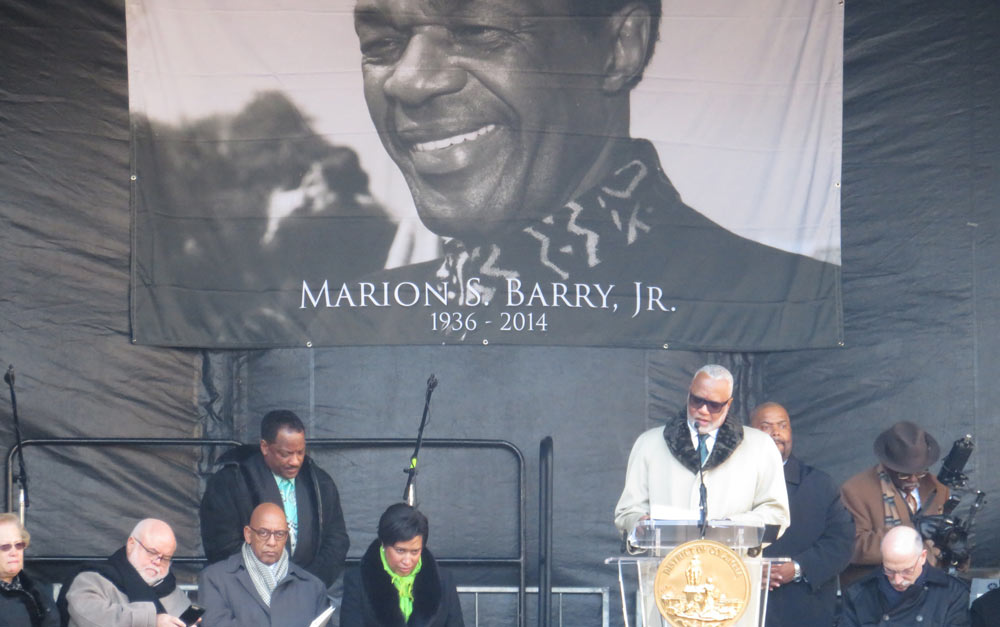Photo of speakers at the podium to dedicate the Marion Barry statue. A giant printed banner of a photograph of Barry hangs in the background.