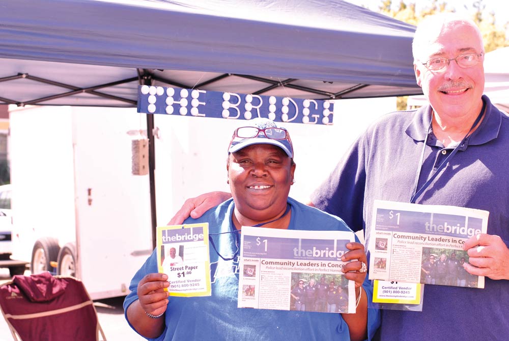 Photo of two people, holding the Memphis Bridge newspaper, with a purple tent and sign saying "The Bridge" in the background.