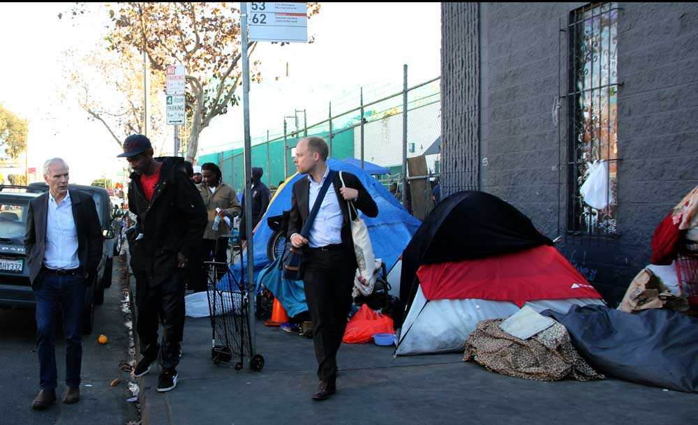 Philip Alston, center, speaks with residents of Skid Row in Los Angeles, California. Tents and debris line the sidewalk behind them. A photographer and assistance follow.