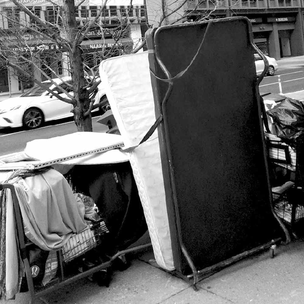 Black and white photo of a bed, cart and other items on a street curb.