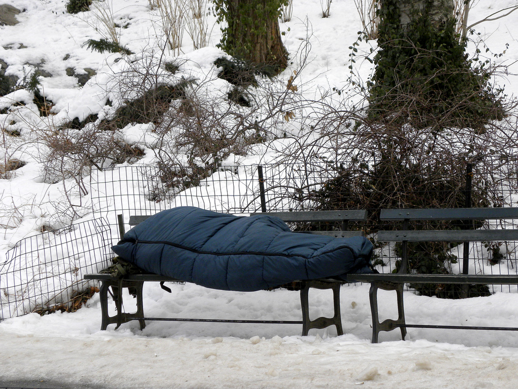 A homeless person lies on a bench in the winter in a sleeping bag