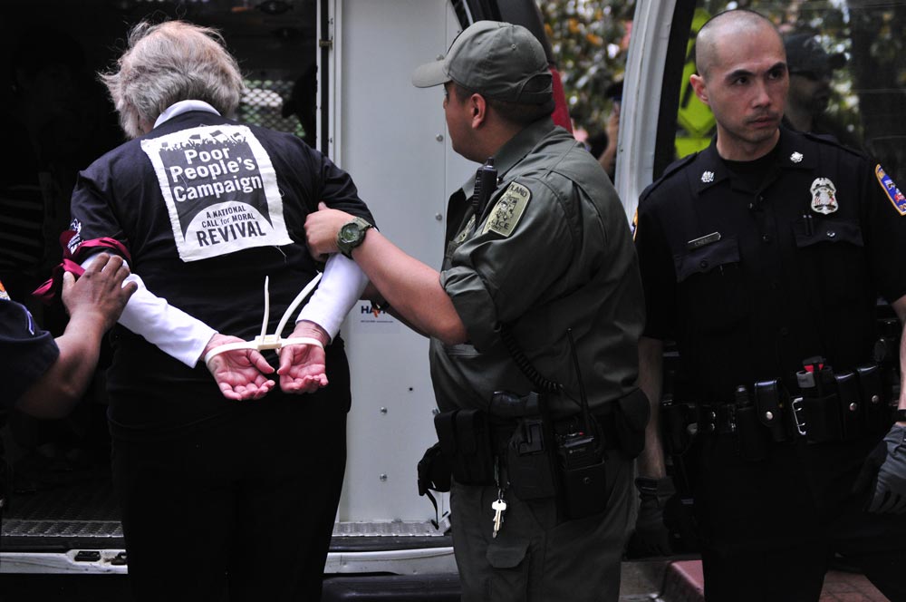 Photo of a woman in zip-tie handcuffs with a Poor People's Campaign sign on her back being led into a police wagon by two officers.