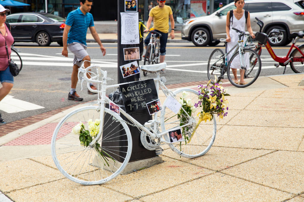 Photo of a white bicycle leaning on a light post with bouquets in the spokes and memorial messages attached to it as well as the light pole.