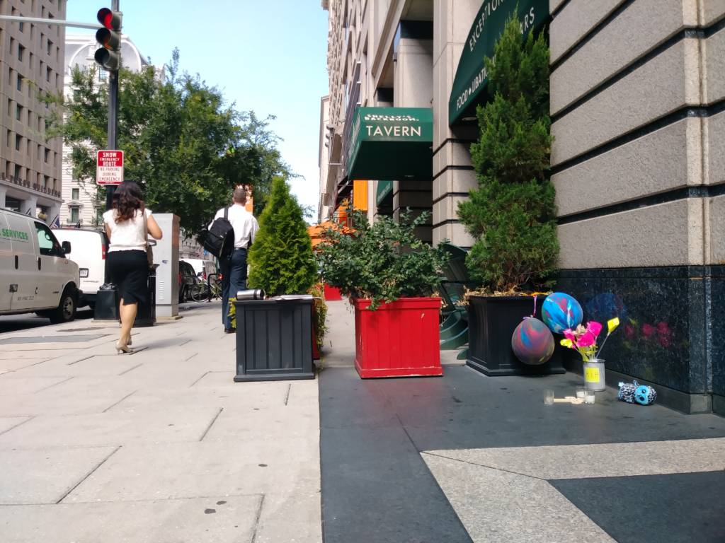 A photo showing balloons, flowers, candles and a cross collected in a corner on the sidewalk. Pedestrians in business attire are walking by.