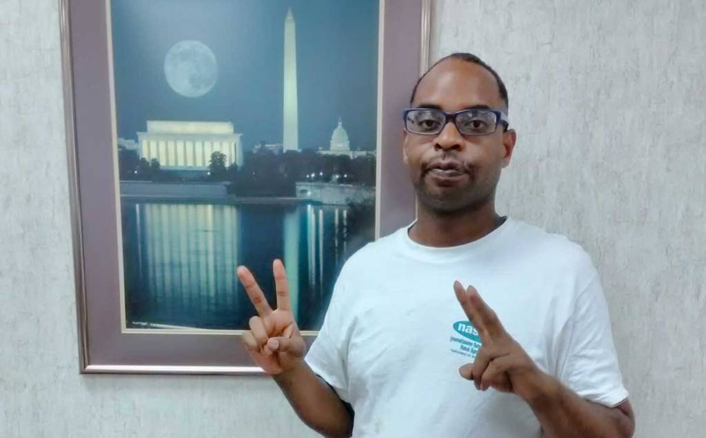 Photo of David displaying two peace signs, one with each hand. He is tanding in front of a picture depicting the Washington and Lincoln monuments at night.
