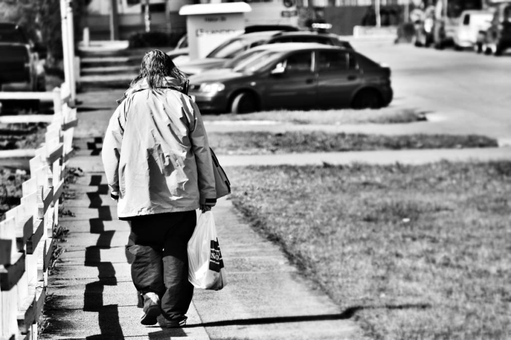 A photograph of a homeless woman walking down a street carrying a shopping bag.