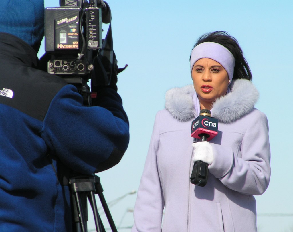 A photograph of a news reporter talking to a news camera being held by a camera operator.