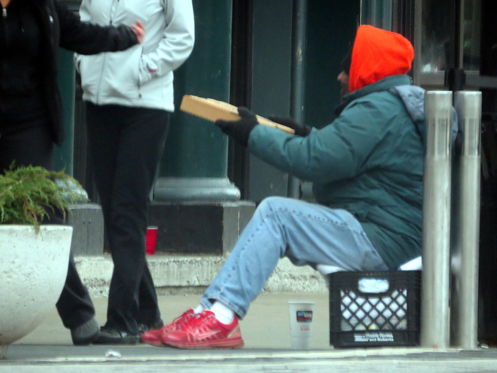 A photograph of a homeless man, bundled against the cold, sitting on a milk crate and holding a pizza box.