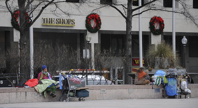 A photograph of a homeless woman, with shopping cart and belongings, sitting in front of a shopping mall displaying Christmas decorations..
