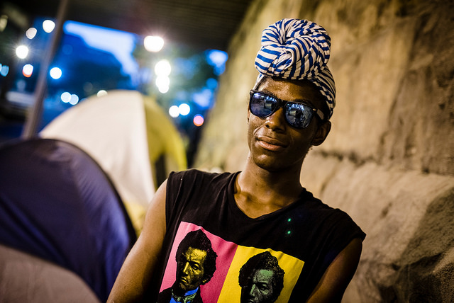 A photograph of a homeless person standing near a stone wall and a tent. It is evening time, and the homeless person is wearing a head-wrap, sunglasses, and a sleeveless T-shirt featuring color images of the young Frederick Douglass