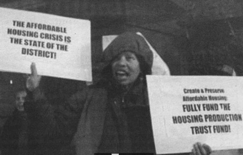 Photo of a woman holding up signs saying: "Create and preserve affordable housing. Fully fund the housing production trust fund!"