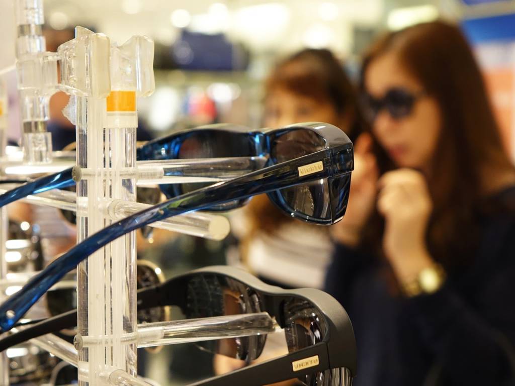Photo of a rack of sunglasses for sale in focus, with two women customers in the background, out of focus.