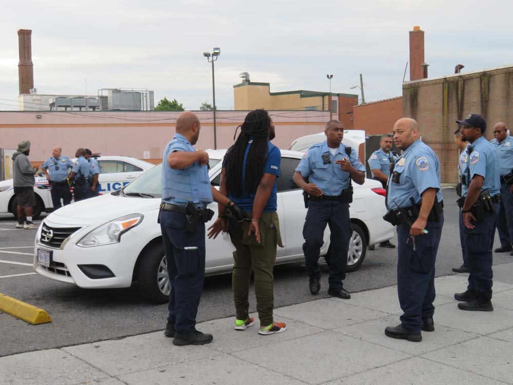 Photo of several police officers around a man standing next to his car. The trunk is popped open.