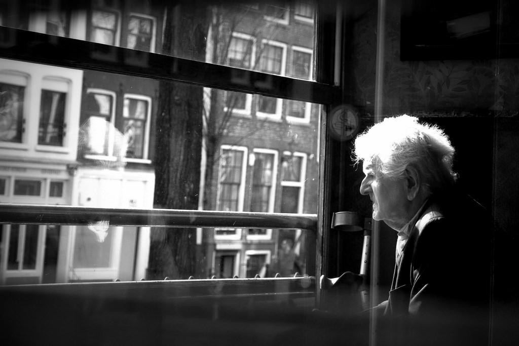 A photograph of an elderly man sitting in a room and looking out of a window.