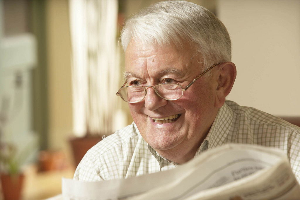 A photograph of an older man, smiling and holding a newspaper.