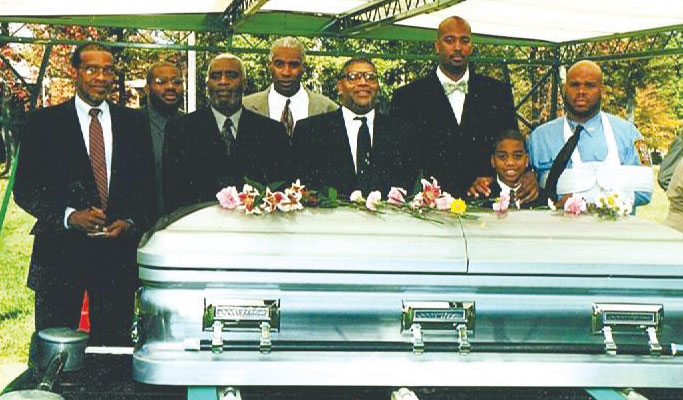 A photograph of Wendell Williams and family, standing aroud his mother's casket at a cemetery.