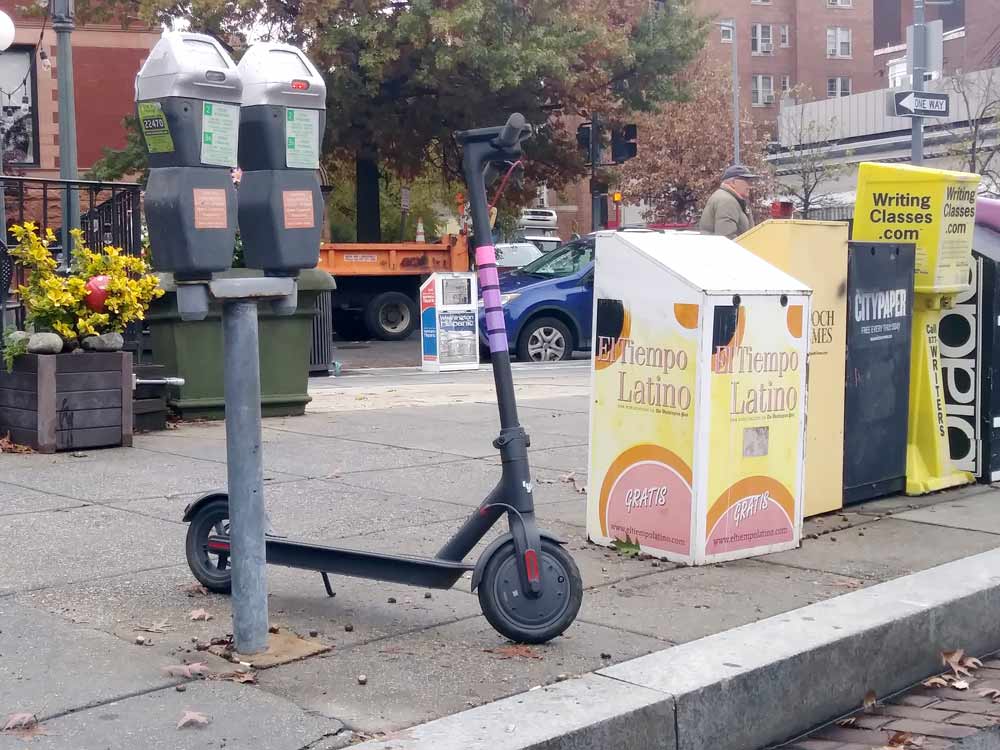 Photo of a Lyft scooter parked between a parking meter and a row of newspaper boxes.