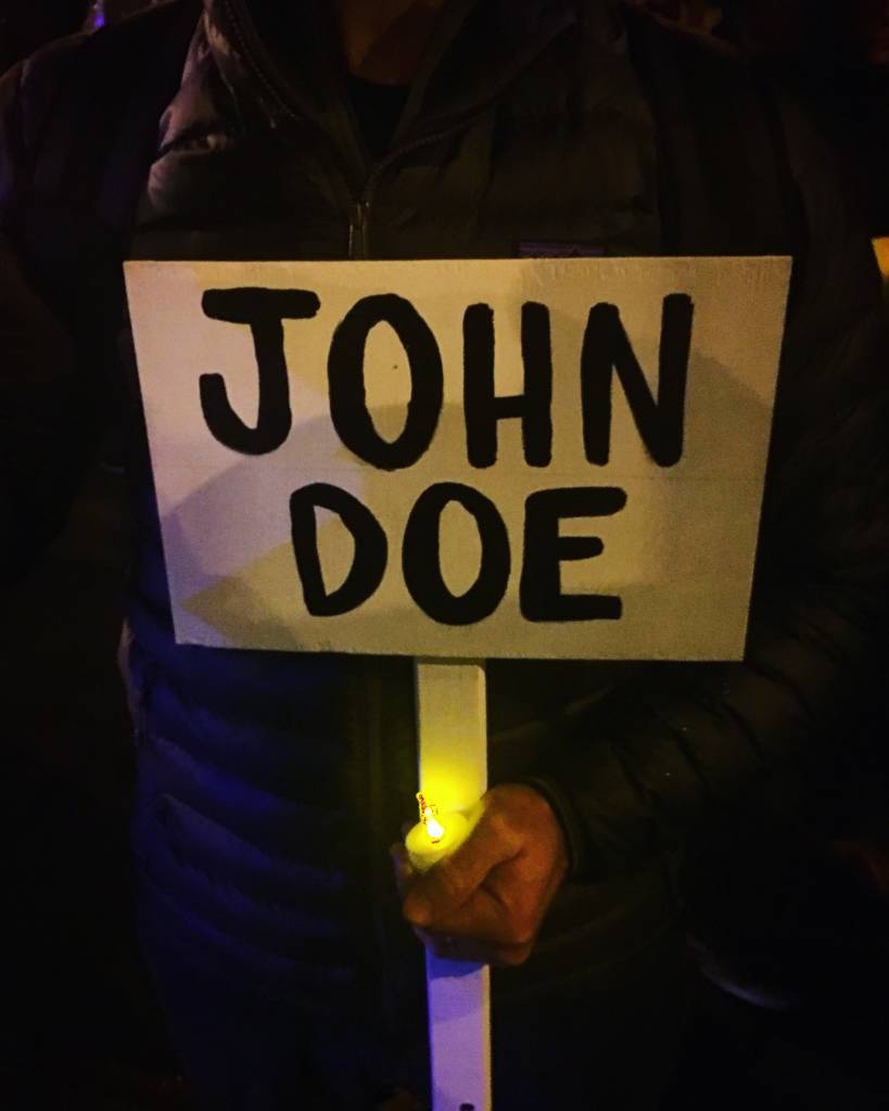 Photo of a white paper sign, attached to a plastic stick. It says "John Doe" and the hands holding it also hold an electric candle.