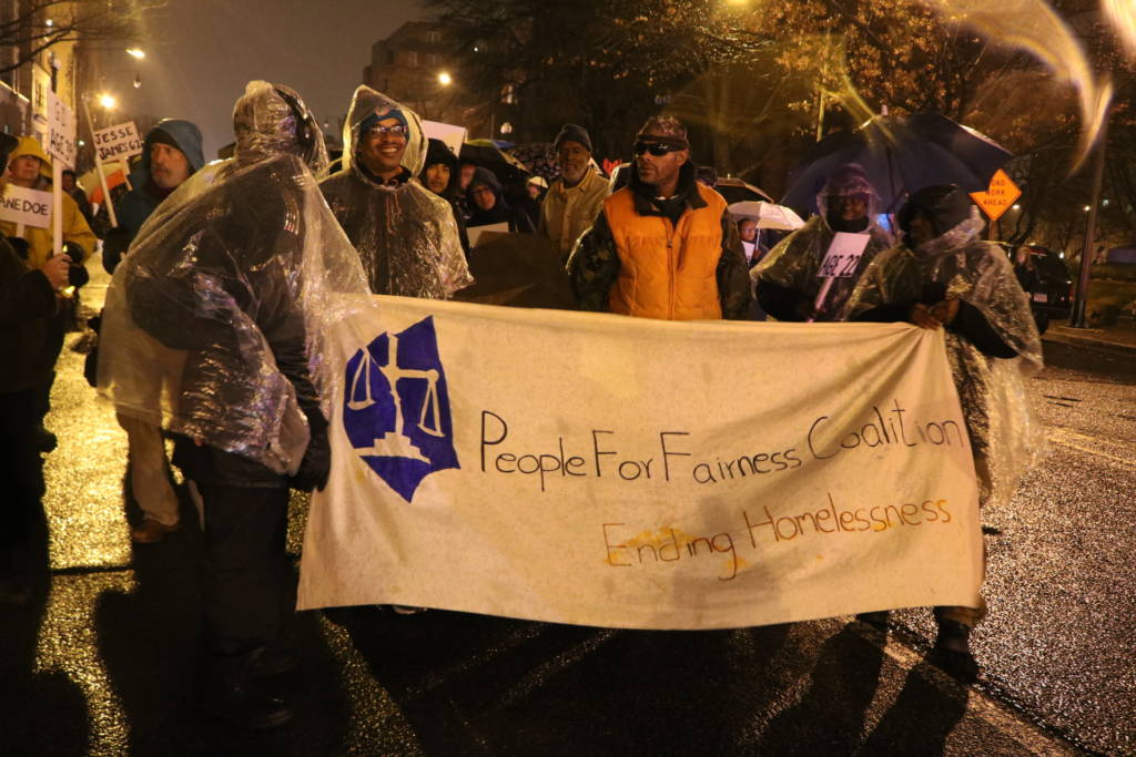 Photo of a group of people walking a t night, holding a banner that says "People for Fairness" linked
